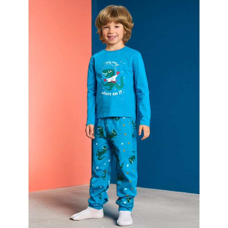 A young boy wearing a bright blue long-sleeve pajama set with a dinosaur-themed graphic on the chest and a matching blue pajama set with green dinosaur prints, standing against a split orange and dark blue studio background.