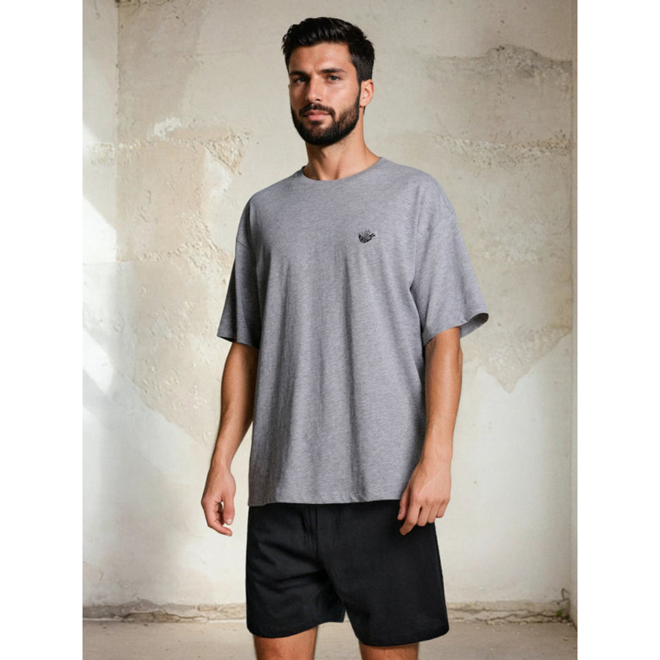 A man wearing an oversized heather gray t-shirt with a small black embroidered logo on the left chest, paired with black shorts, standing against a textured concrete wall.