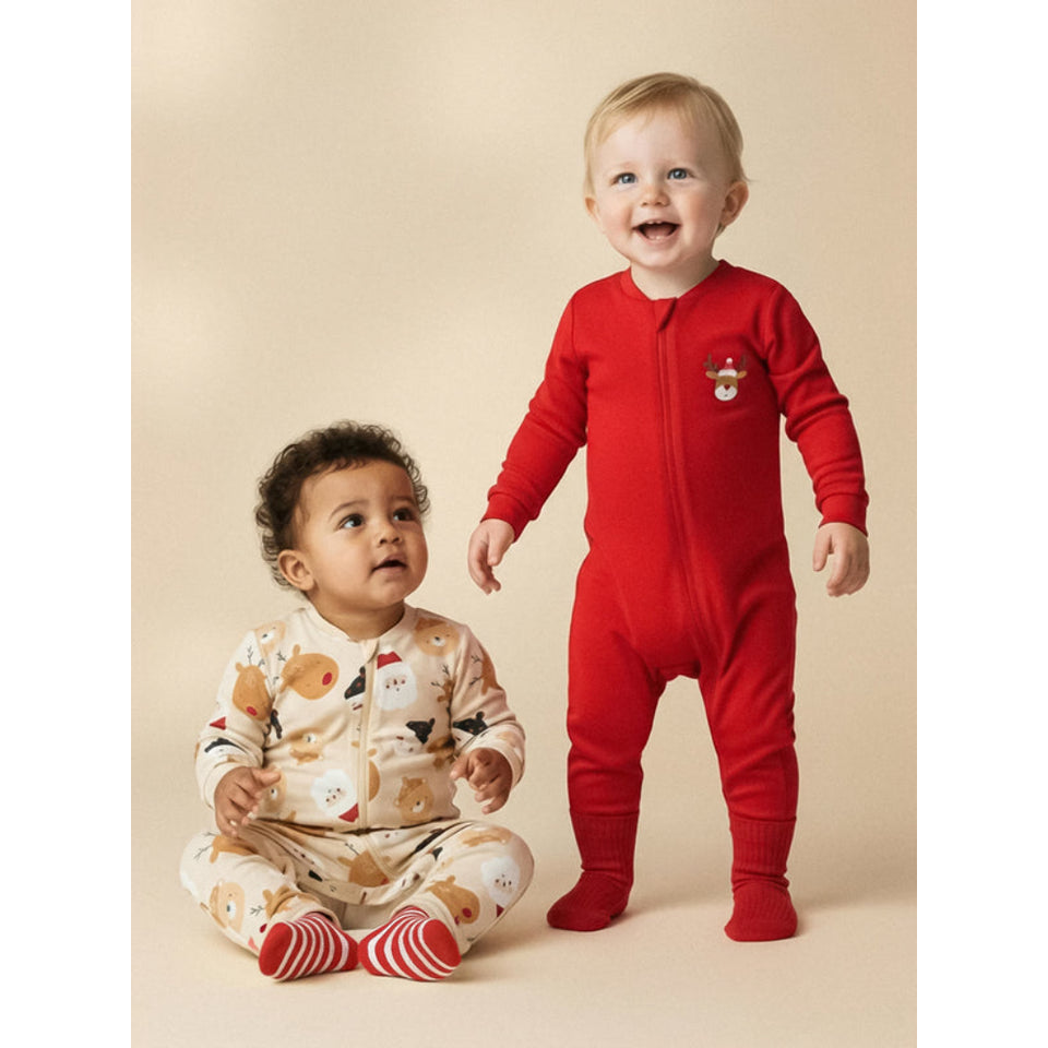 Two toddlers wearing holiday footed pajamas pose against a warm beige studio backdrop. The standing toddler wears a bright red one-piece pajama with a small reindeer chest detail, while the seated toddler wears a cream pajama with a teddy bear pattern and red-and-white striped socks.