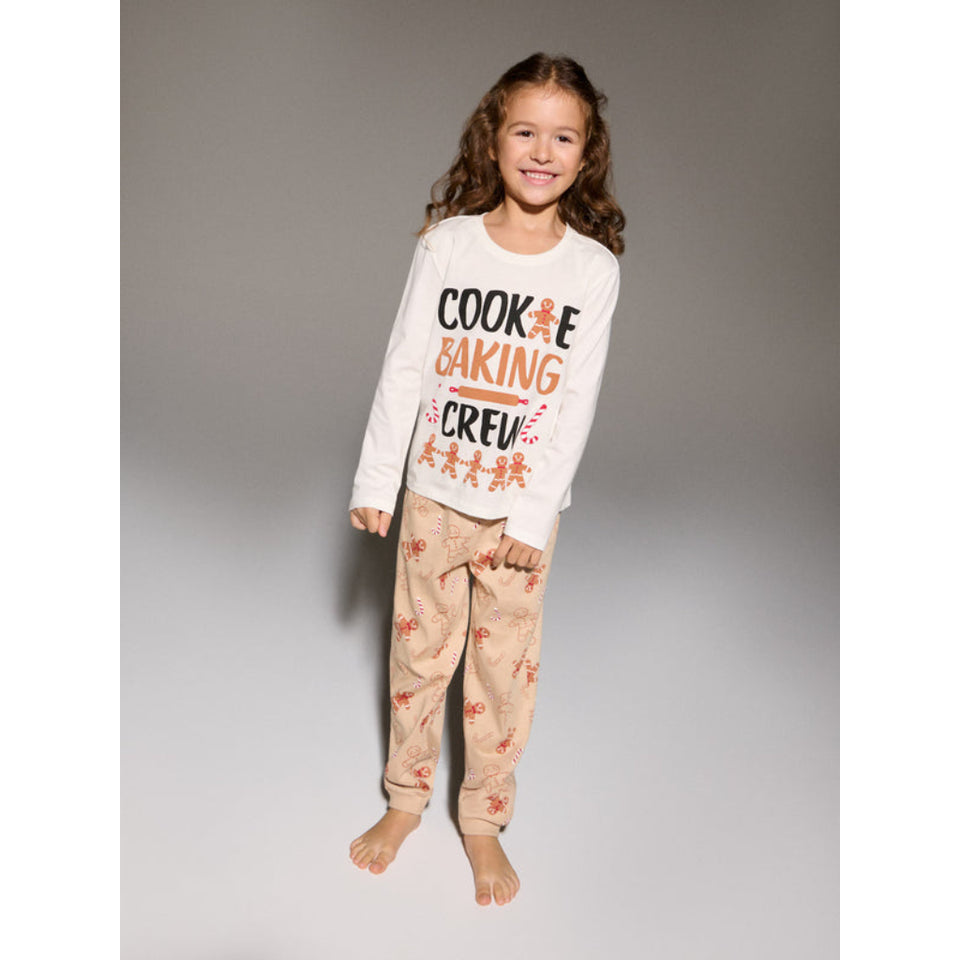A young girl stands barefoot against a neutral gray studio background, wearing a white long-sleeve pajama top with the words 'COOKIE BAKING CREW' in black and orange, paired with beige pants featuring a small orange cookie print.
