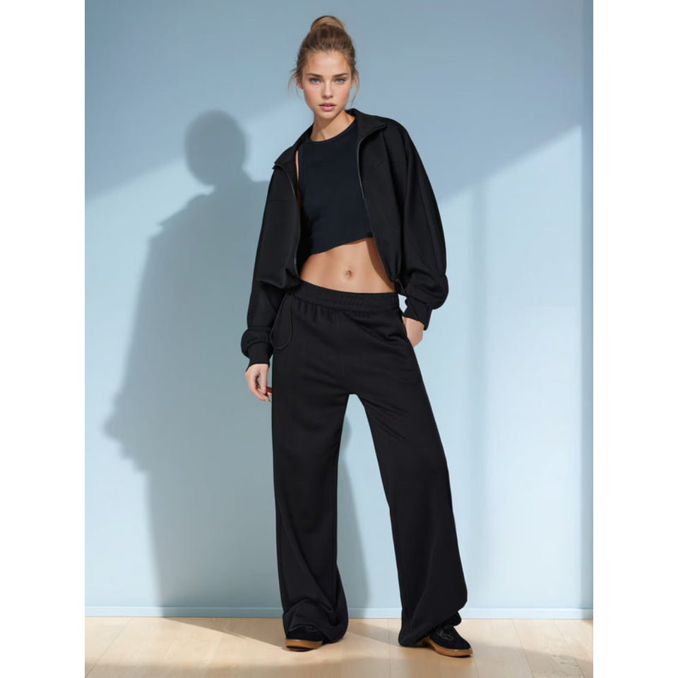 A model stands against a light blue wall wearing a black cropped tank top, a black zip-up hoodie draped off the shoulders, and black wide-leg sweatpants.