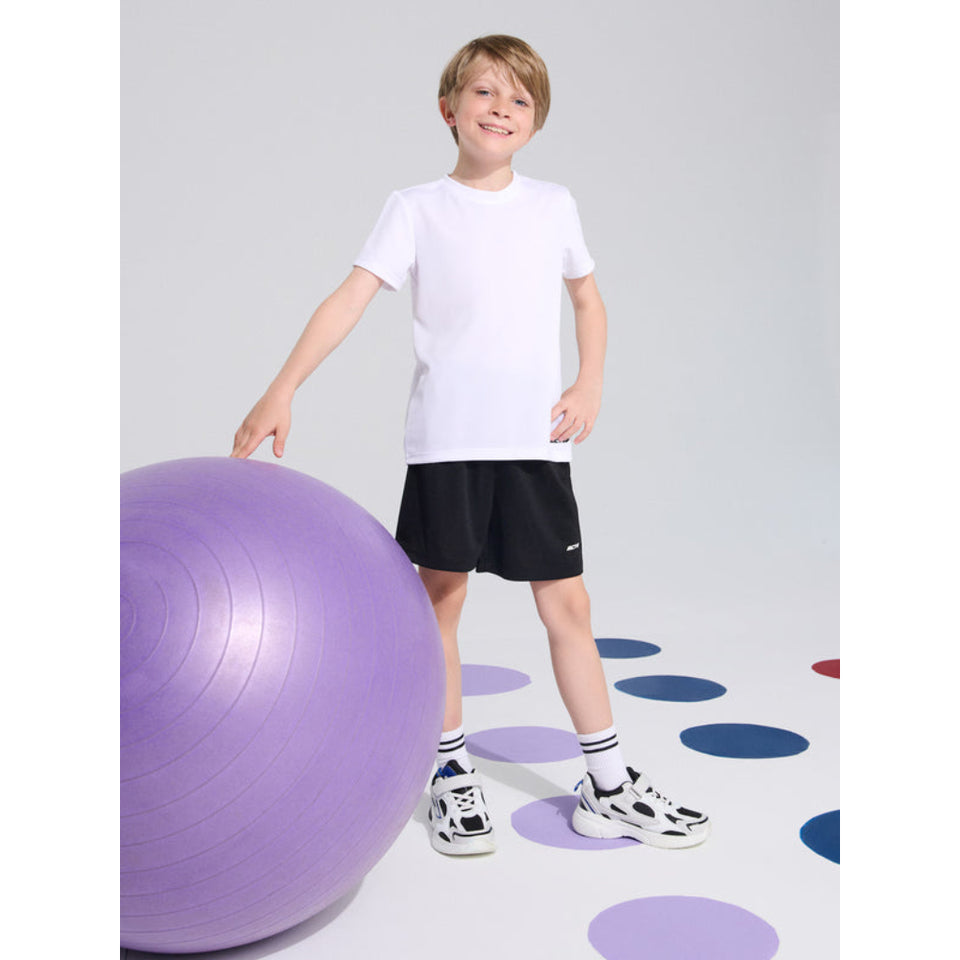 A young boy wearing a white short-sleeve t-shirt, black athletic shorts, and white sneakers stands next to a large purple exercise ball in a studio setting with a light gray background and colorful polka dots on the floor.