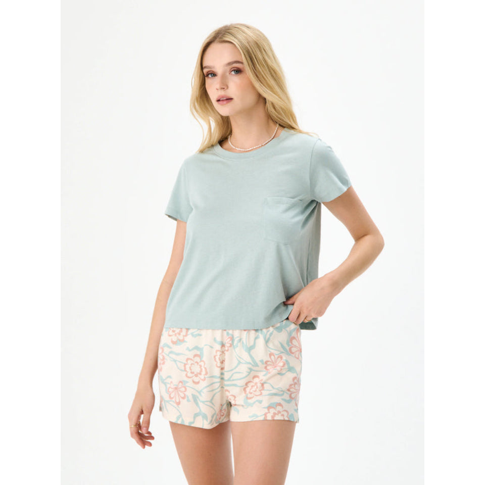 A woman modeling a light sage green short-sleeve crewneck t-shirt with a chest pocket, paired with cream floral shorts, against a plain white studio background.