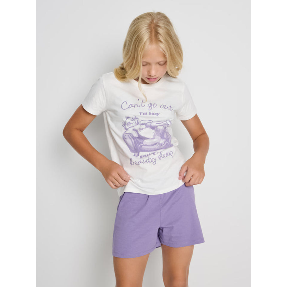 A young girl with blonde hair is wearing a white graphic t-shirt and lavender purple shorts against a light gray studio background.