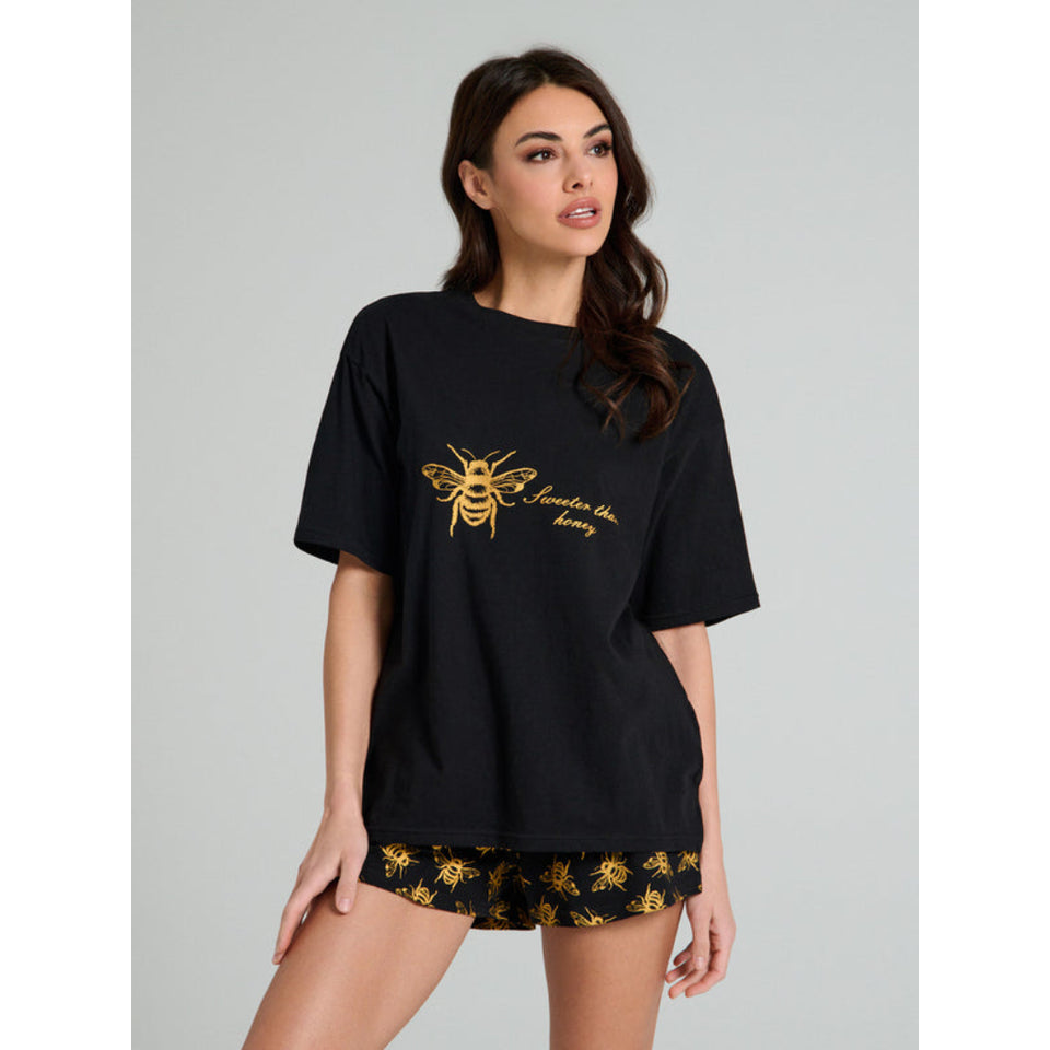 A woman modeling an oversized black t-shirt with a gold bee graphic and the text 'Jamboree' on the chest, paired with black shorts featuring a gold bee pattern, against a solid gray studio background.