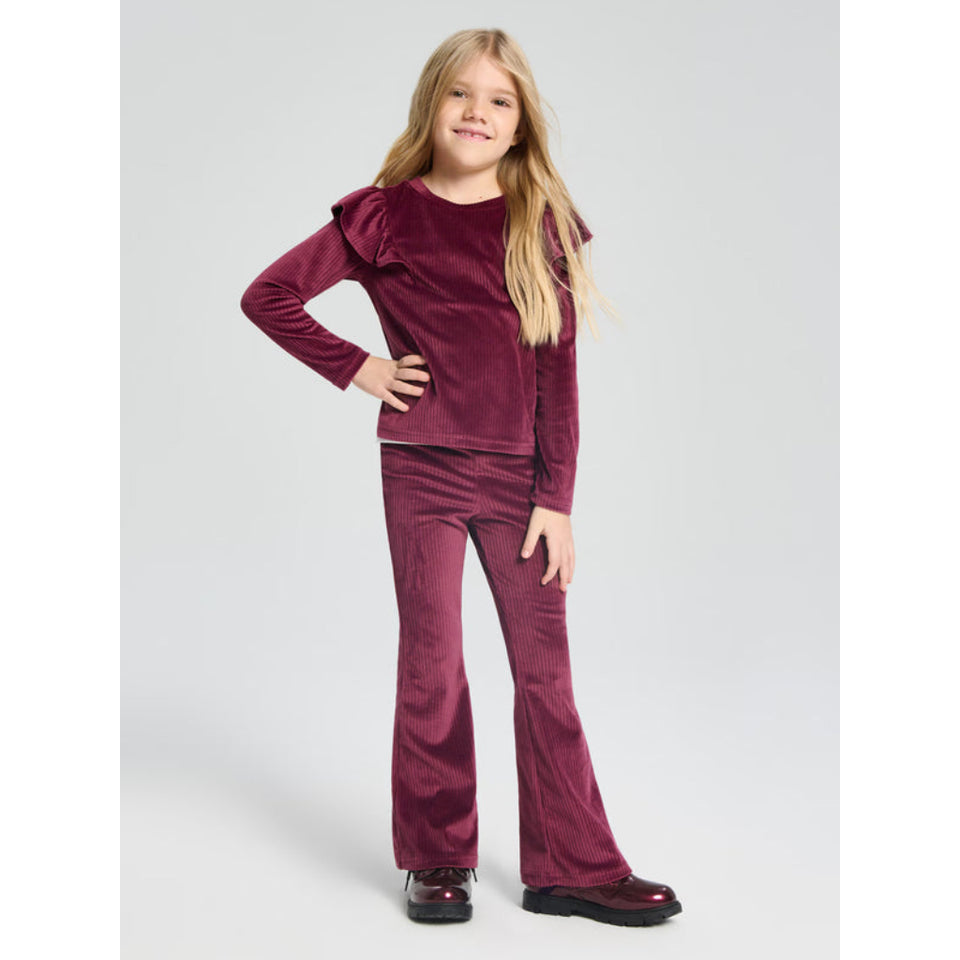 A young girl with blonde hair stands against a light gray studio background wearing a matching burgundy velvet long-sleeve top with ruffle details on the shoulders and matching flared pants.