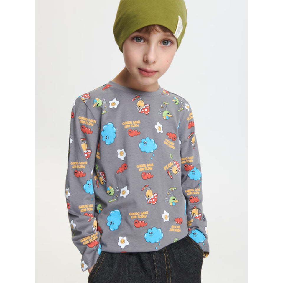 A child wearing a gray long-sleeve crewneck sweatshirt with a colorful cartoon cloud and egg pattern, paired with a green beanie and dark jeans, against a light gray studio background.