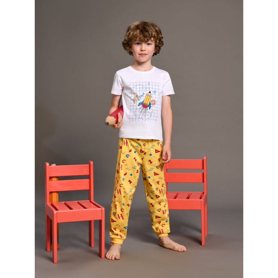 A young boy stands barefoot between two coral-red chairs against a plain gray studio background, wearing a white graphic T-shirt and yellow patterned pajama pants. He holds a small plush toy.