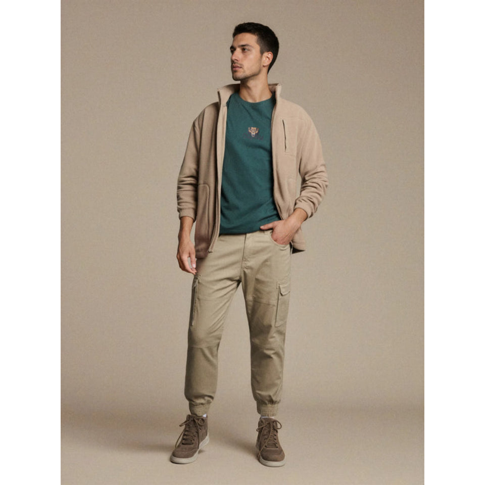 A man modeling a beige zip-up hoodie over a dark teal t-shirt, paired with beige cargo pants and brown sneakers, against a solid beige studio background.