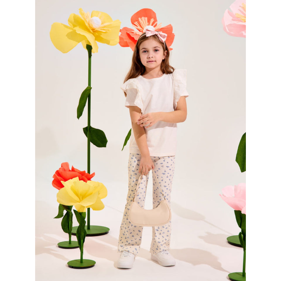 A young girl stands in a studio wearing a white short-sleeve top, cream pants with small blue floral prints, white sneakers, and holding a cream crescent-shaped handbag. She is wearing a large coral-orange flower headband.