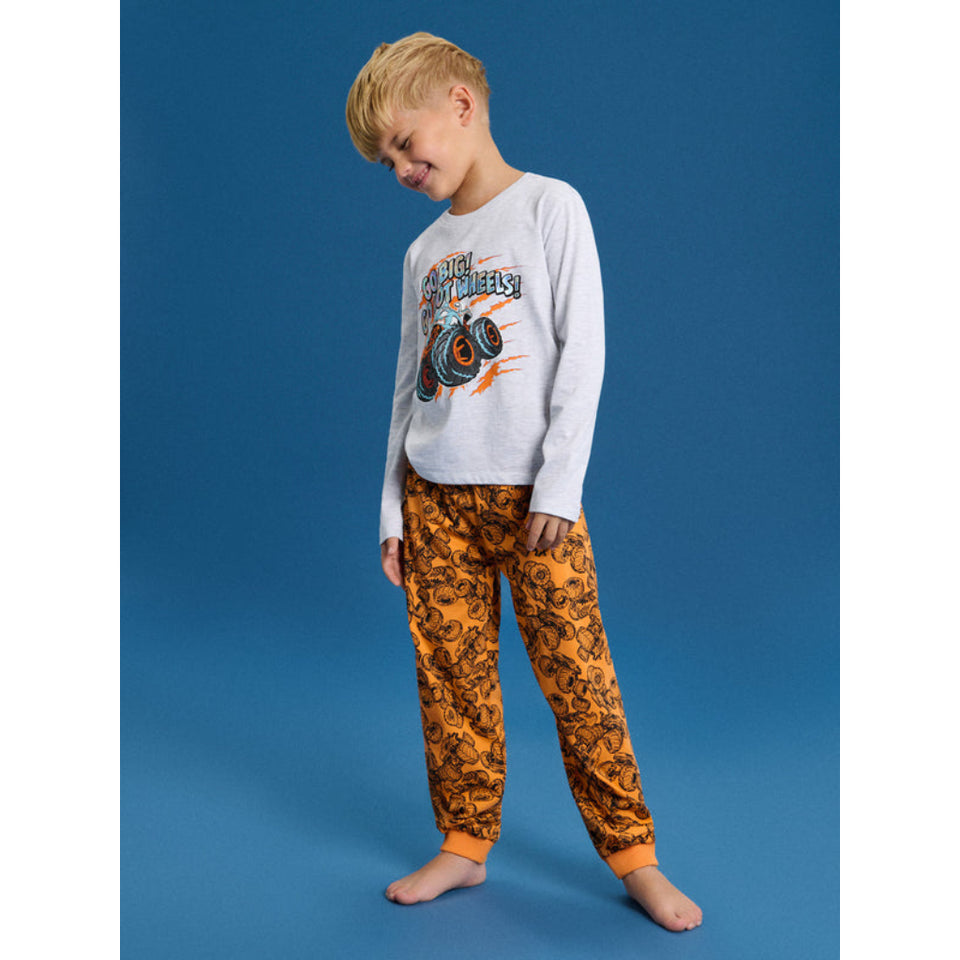 A young boy wearing a two-piece pajama set consisting of a light gray long-sleeve shirt with a monster truck graphic and orange patterned pants, standing against a solid blue studio background.