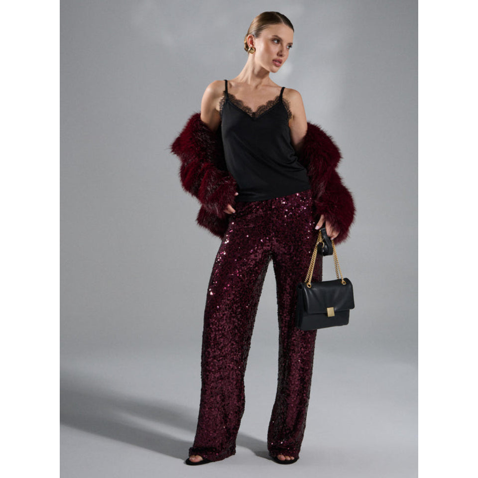 A model stands against a gray studio background wearing a black camisole, wide-leg sequined trousers in deep burgundy, a matching burgundy faux-fur jacket draped off the shoulders, and holding a black handbag with gold hardware.