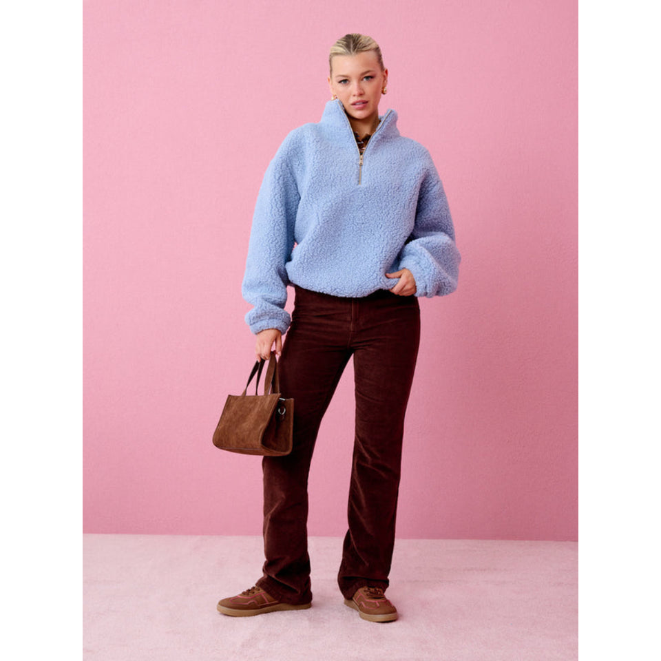 A model stands against a pink studio backdrop wearing a light blue fuzzy quarter-zip pullover, dark brown corduroy pants, and brown shoes, while holding a brown handbag.