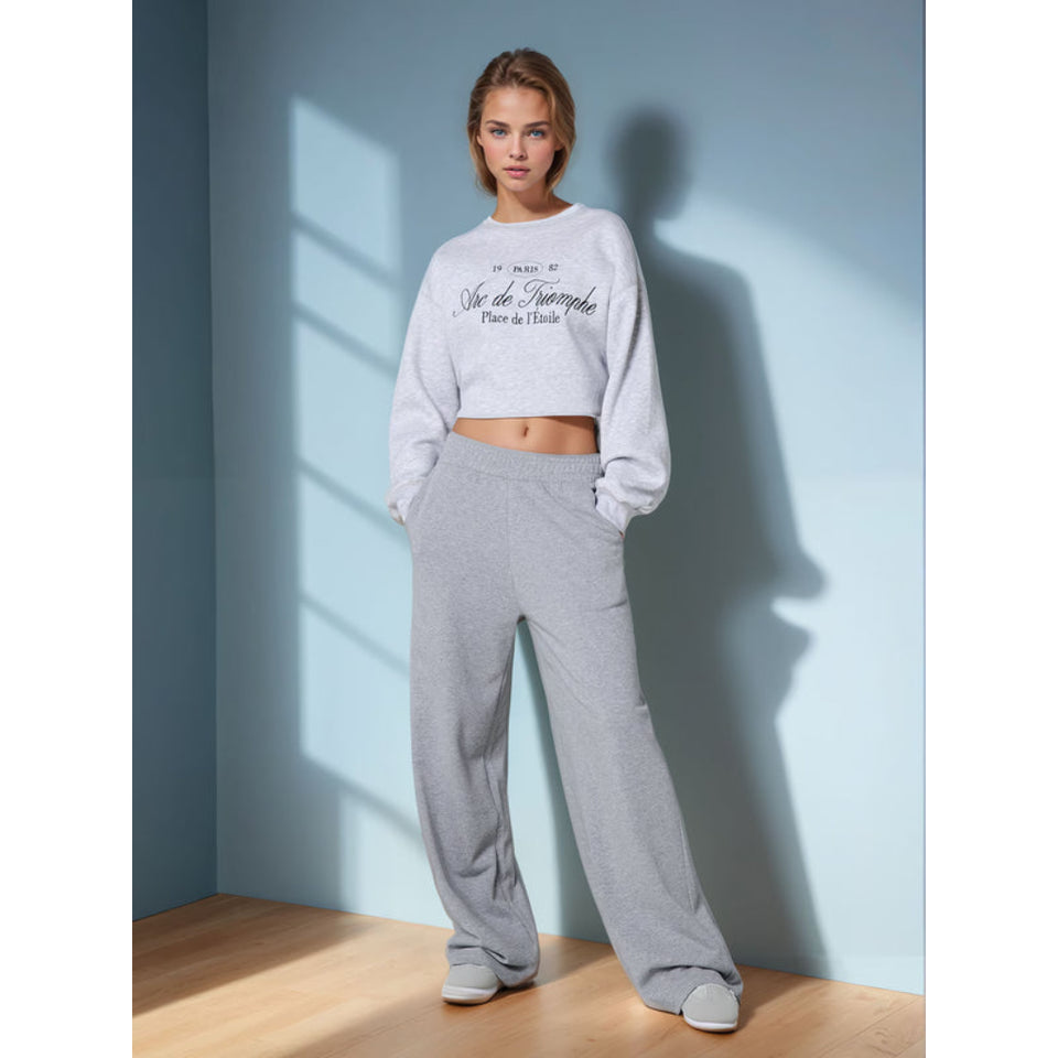 A woman modeling a light gray cropped sweatshirt and matching wide-leg sweatpants in a studio setting with a blue wall and wooden floor.