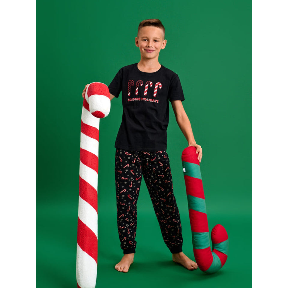 A young boy stands barefoot against a solid green studio background, holding two oversized candy cane plush toys. He is wearing a black graphic t-shirt and matching patterned pajama pants.