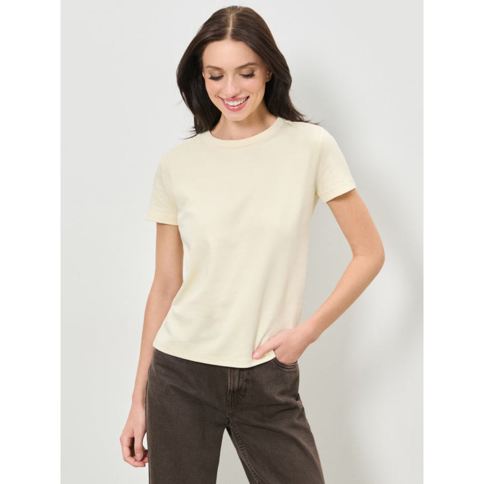 A woman modeling a short-sleeve crewneck t-shirt in a light cream color, paired with dark brown pants, against a light gray studio background.