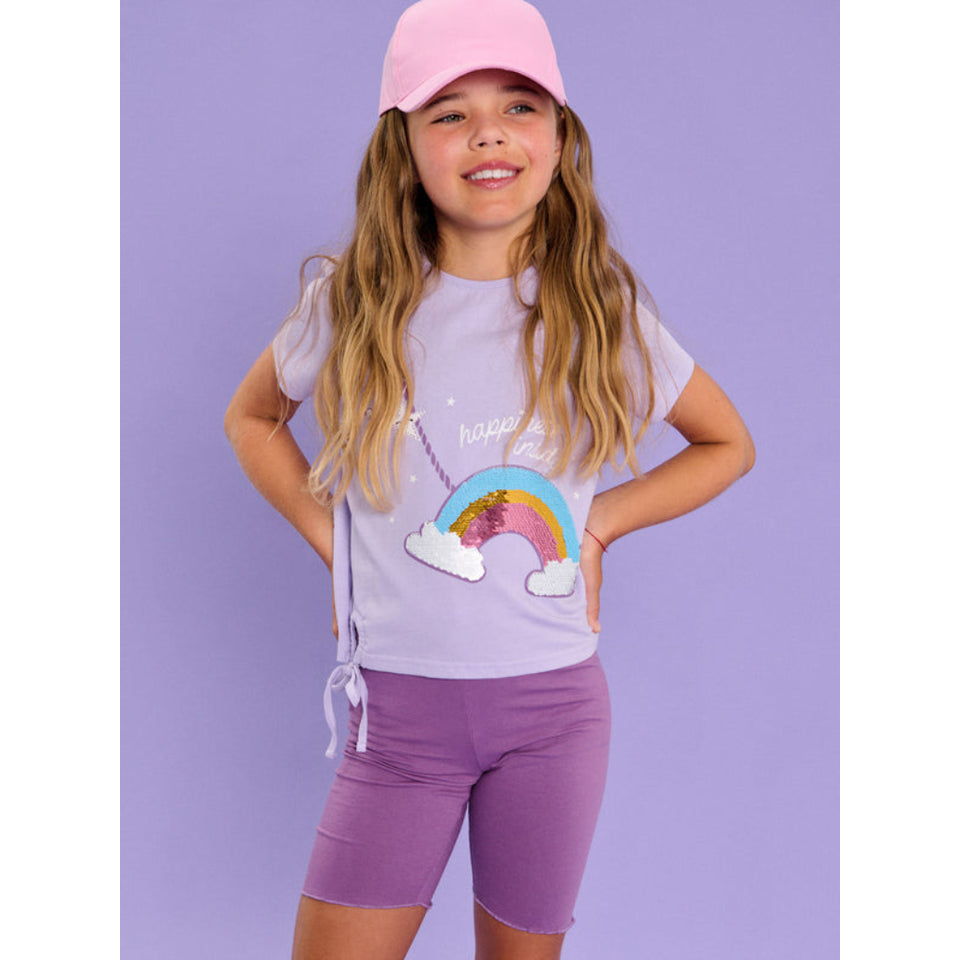 A young girl modeling a light purple t-shirt with a rainbow sequin graphic, matching purple biker shorts, and a pink baseball cap, set against a solid lavender background.