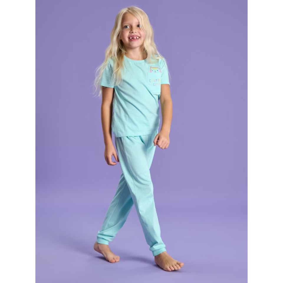 A young girl with long blonde hair stands barefoot against a solid lavender studio background, wearing a light mint green two-piece pajama set with a small rainbow graphic on the chest.