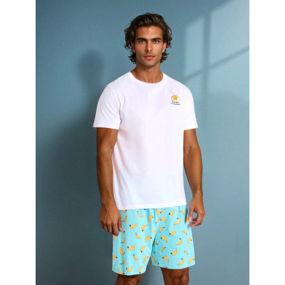 A man modeling a white short-sleeve t-shirt with a small yellow and blue logo on the left chest, paired with light blue shorts featuring a yellow banana print, set against a blue-gray wall.