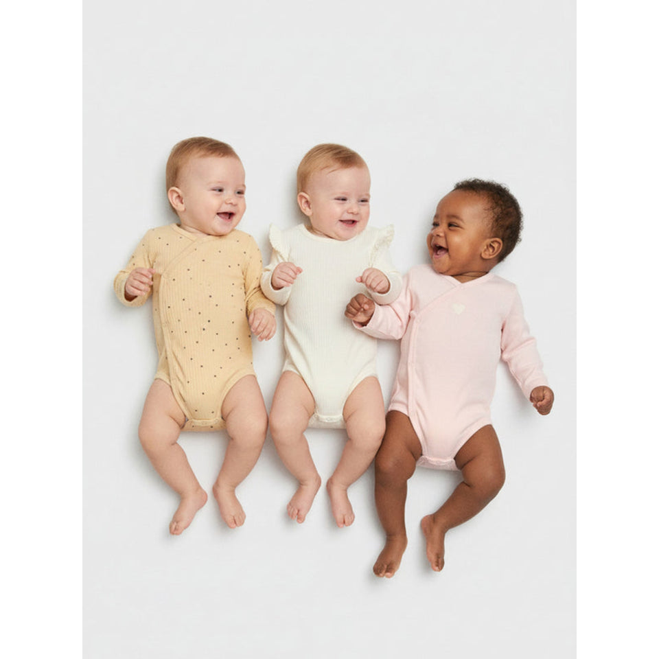Three babies lying side by side on a light gray studio background, each wearing a long-sleeve one-piece bodysuit in a different soft pastel color: pale yellow with tiny dark dots, cream, and light pink.