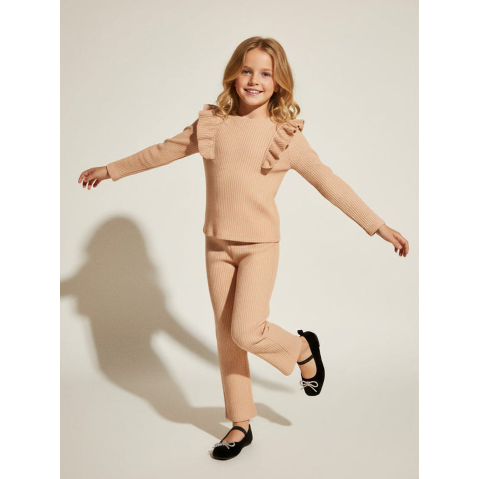 A young girl with blonde hair is wearing a beige ribbed knit two-piece outfit consisting of a long-sleeve top with ruffle details on the shoulders and matching pants. She is posing with her arms outstretched on a light beige studio background, casting a shadow.