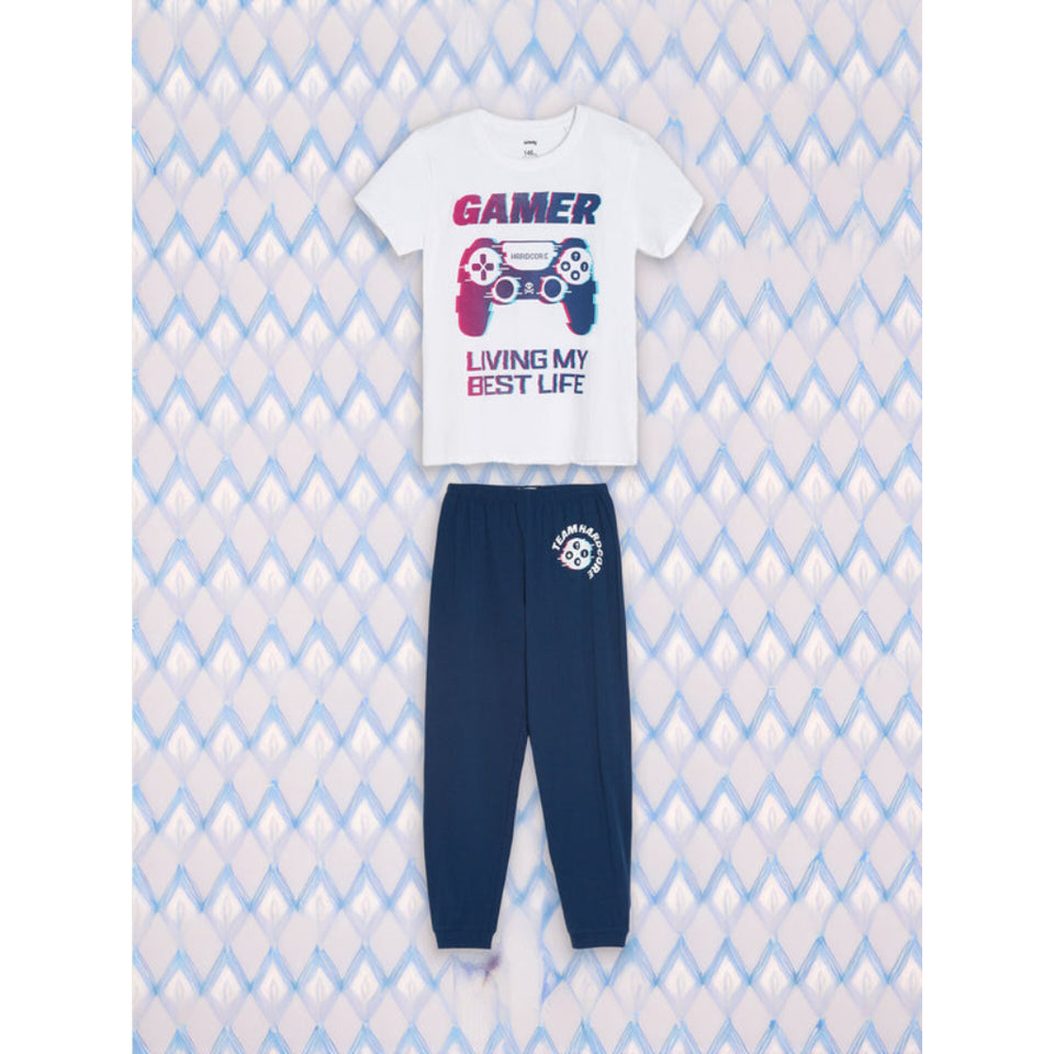 A two-piece kids' pajama set featuring a white short-sleeve t-shirt with a colorful video game controller graphic and the text 'GAMER LIVING MY BEST LIFE' in blue and pink, paired with navy blue jogger pants with a white circular logo on the left thigh, laid flat against a light blue and white patterned background.