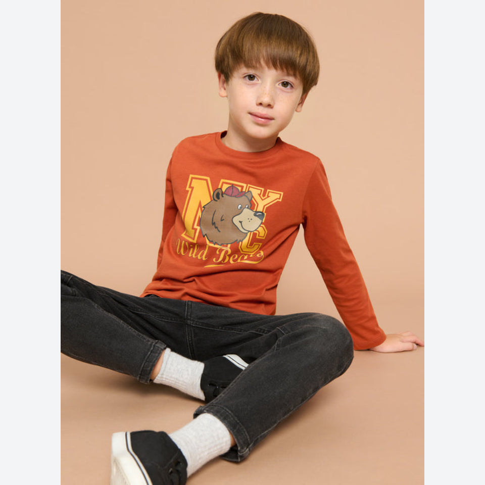 A young boy sitting on a beige studio floor wearing a rust-orange long-sleeve graphic t-shirt with a cartoon beaver and the text 'Beaver Jazz', paired with black jeans, white socks, and black sneakers.