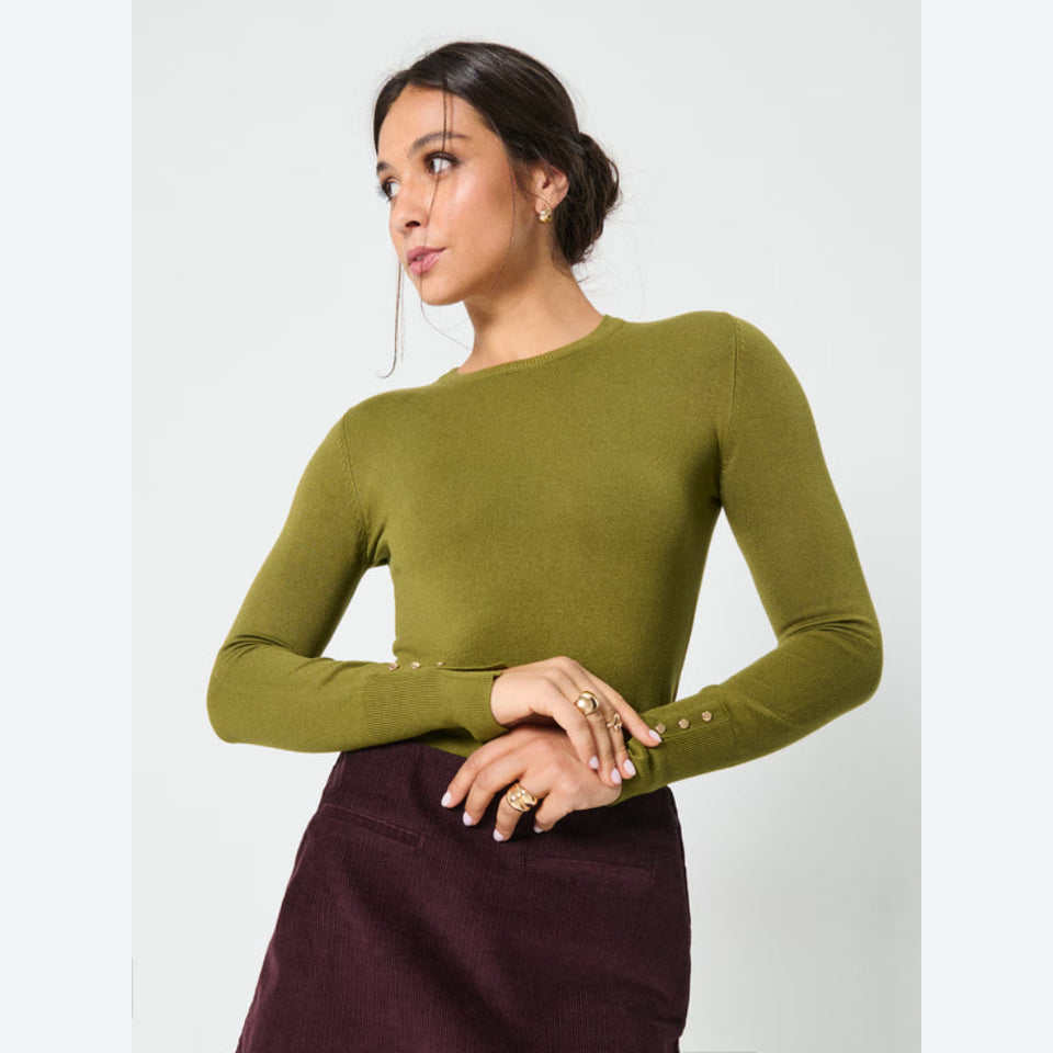 A woman wearing a fitted olive green long-sleeve knit top with decorative gold buttons along the cuffs and waist, paired with a dark brown skirt, against a light gray studio background.