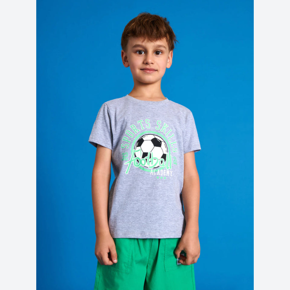 A young boy wearing a heather gray t-shirt with a green and black soccer graphic that reads 'Soccer Academy Football', paired with green shorts, standing against a solid blue background.