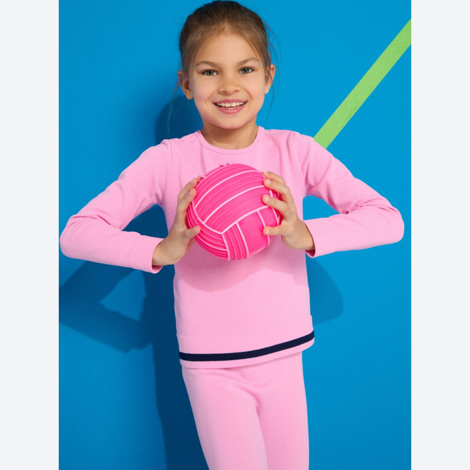 A young girl smiling and holding a pink volleyball, wearing a matching pink long-sleeve athletic top and leggings with a thin black stripe at the hem, posed against a bright blue background with a lime green diagonal line.