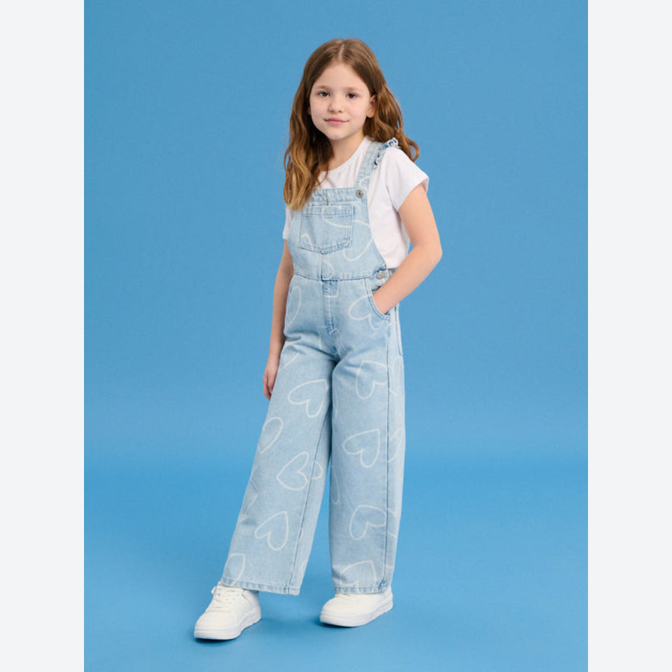 A young girl modeling light blue denim overalls with a white heart pattern, worn over a white t-shirt and white sneakers, set against a solid blue studio background.