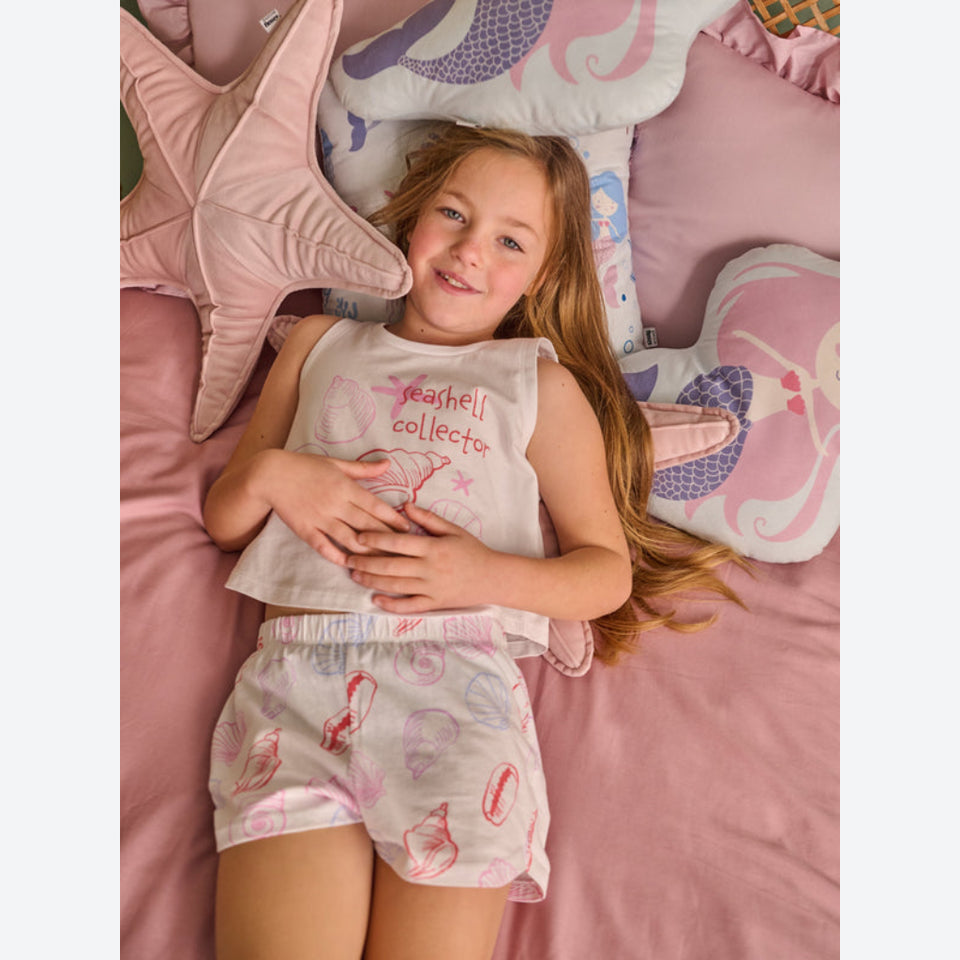 A young girl lies on a pink bed wearing a matching sleeveless top and shorts set with a seashell pattern and the words 'teacher collector' printed on the front.
