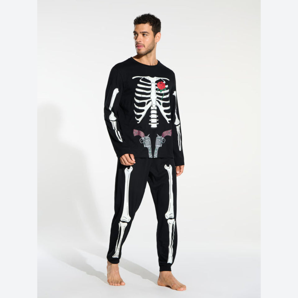 A man modeling a black long-sleeve pajama top and matching pants set with a white skeleton print. The top features a white rib cage graphic with a small red rose at the center chest, and the pants have white bone graphics down the legs.
