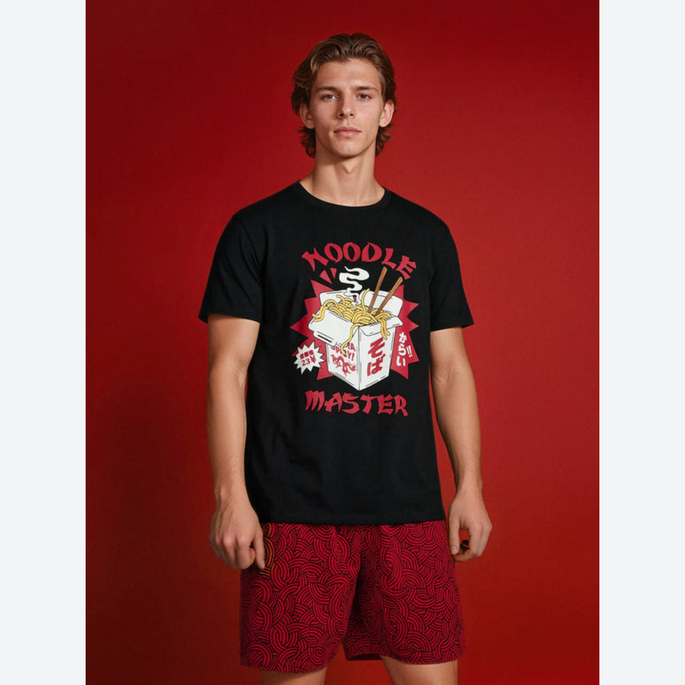 A man modeling a black graphic t-shirt with a red, white, and yellow noodle box design that reads 'NOODLE MASTER', paired with red patterned shorts, set against a solid red studio background.