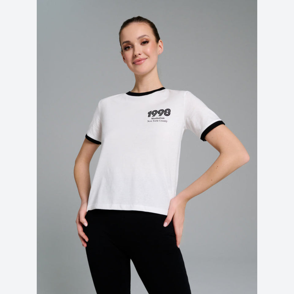 A woman modeling a white ringer T-shirt with black trim at the crew neck and sleeve cuffs, featuring a black '1998' graphic on the left chest. She is wearing black leggings against a solid gray studio background.