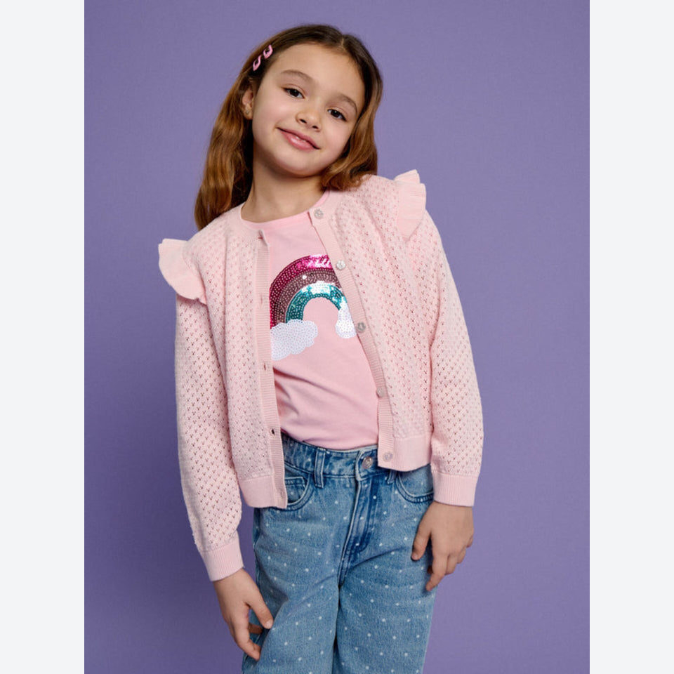 A young girl wearing a light pink knit cardigan with ruffle details, a pink sequined rainbow graphic t-shirt, and blue jeans, standing against a solid purple studio background.