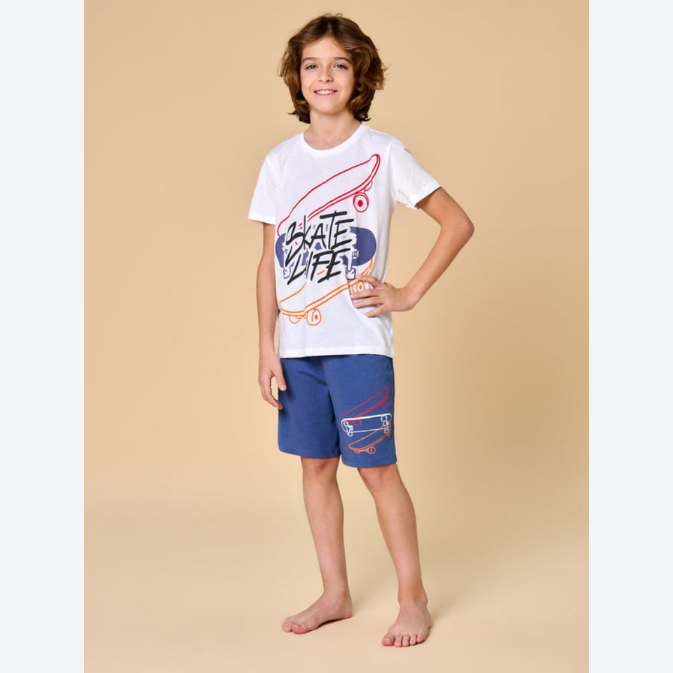 A young boy stands barefoot against a solid beige studio background, wearing a white graphic t-shirt with a skateboard design and blue shorts with a skateboard print on the leg.