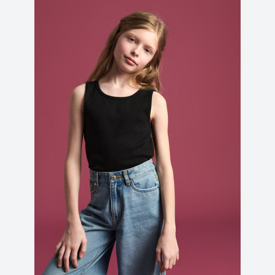 A young girl with light brown hair wearing a black sleeveless tank top and light blue jeans, posing against a solid dusty rose background.