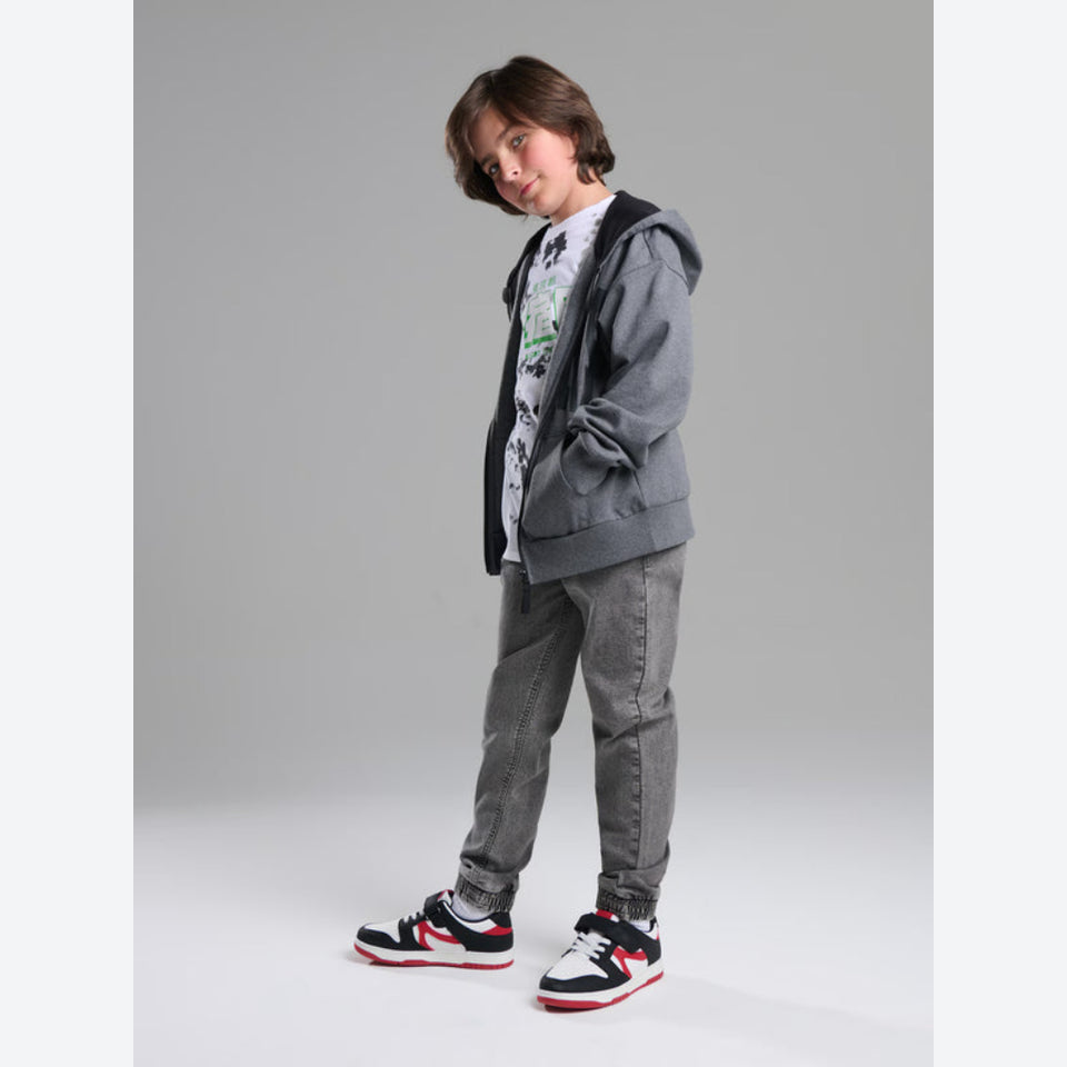 A child model wearing a gray zip-up hoodie over a white graphic t-shirt, gray jeans, and black, white, and red sneakers, standing against a solid gray studio background.