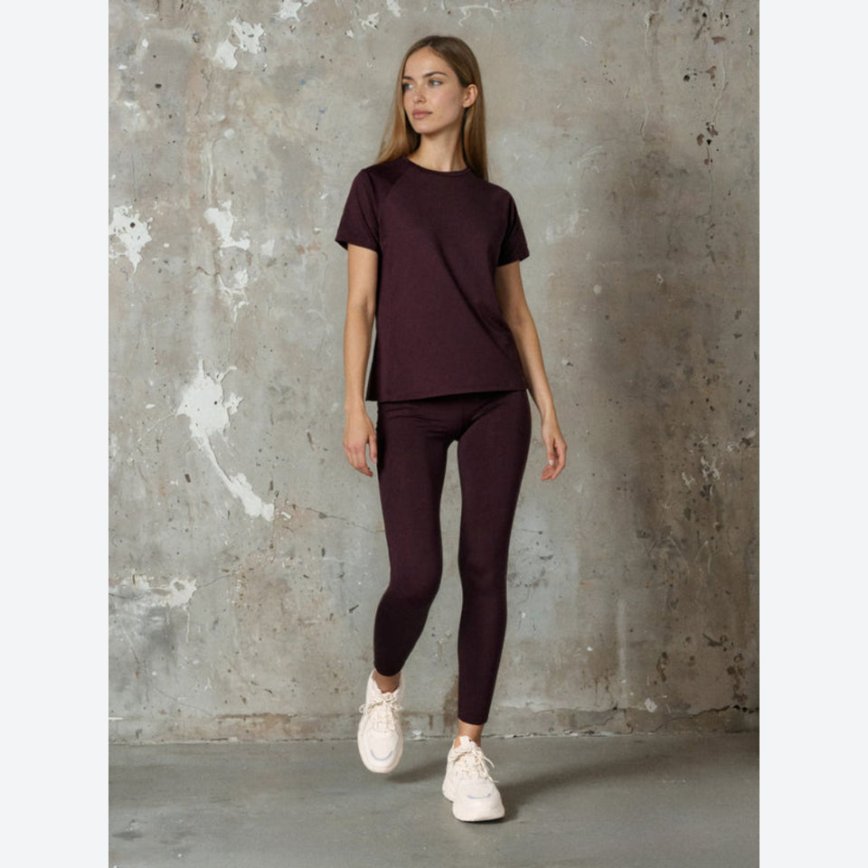 A woman modeling a matching dark plum purple activewear set consisting of a short-sleeve crewneck t-shirt and high-waisted leggings, paired with white sneakers, standing against a textured concrete wall.