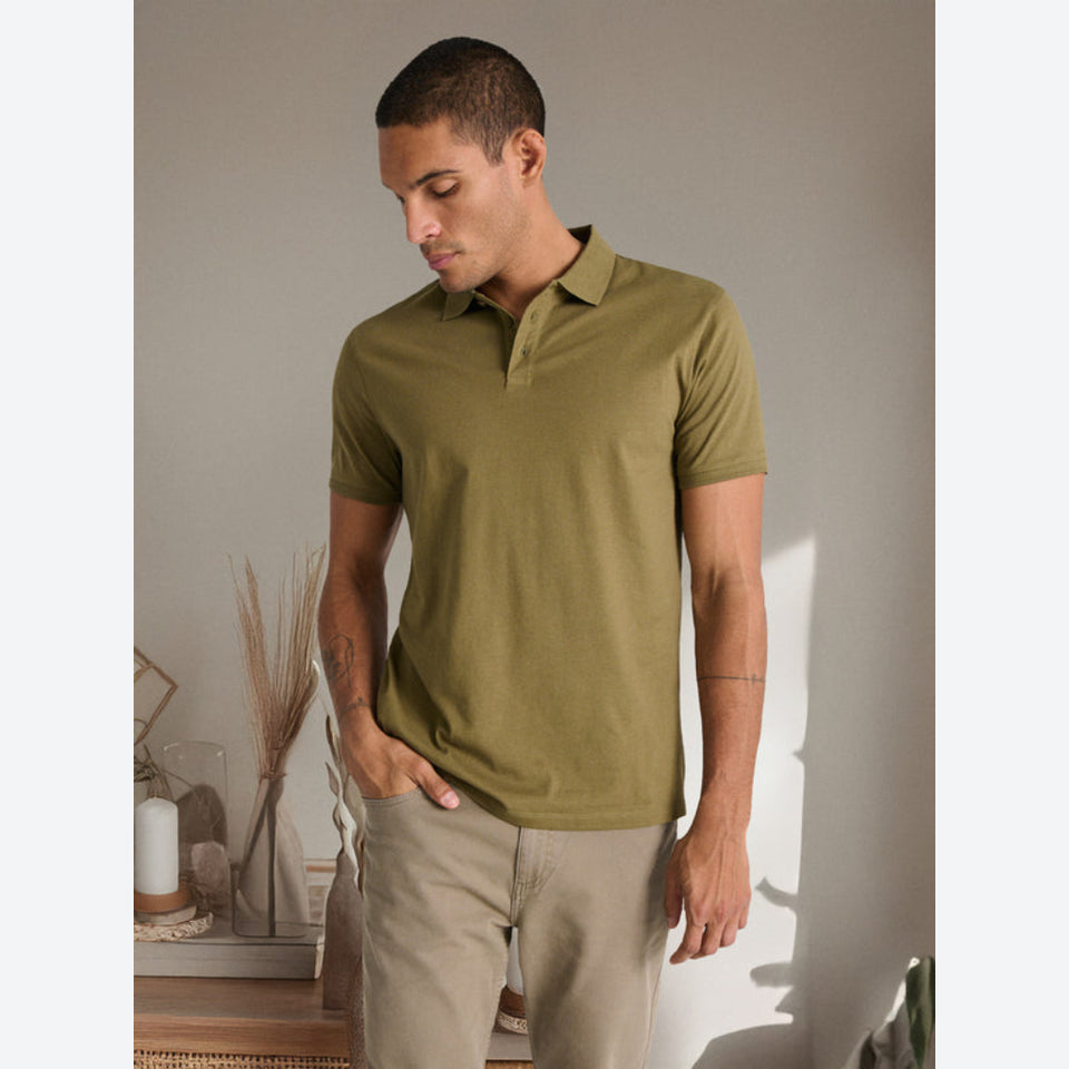A man wearing a short-sleeve olive green polo shirt and beige trousers, standing in a softly lit indoor setting with a neutral wall and decorative dried grasses in the background.