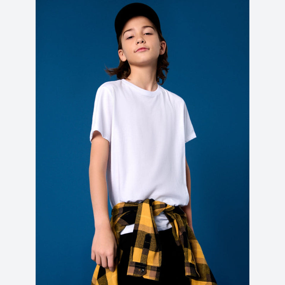 A child model wearing a plain white short-sleeve crewneck t-shirt, a black baseball cap, and a yellow and black plaid flannel shirt tied around the waist, set against a solid blue studio background.