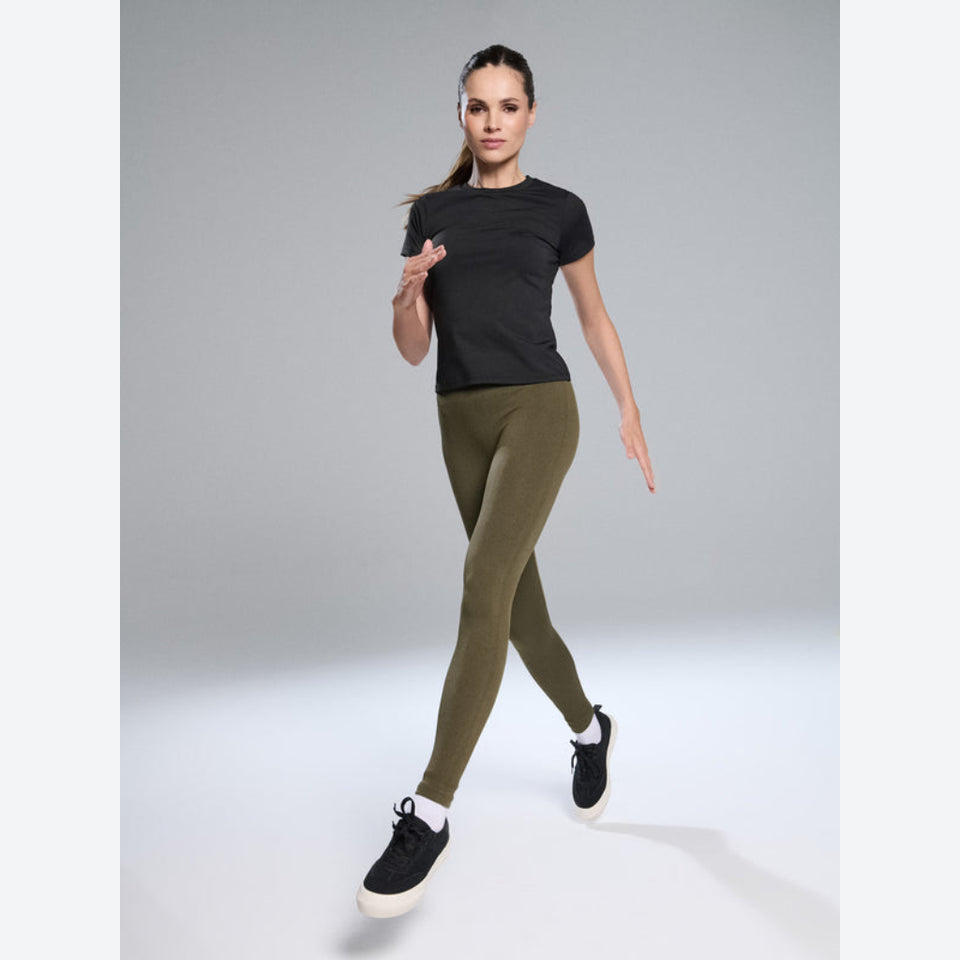 A woman modeling a black short-sleeve athletic top and olive green leggings with black sneakers, posed against a light gray studio background.