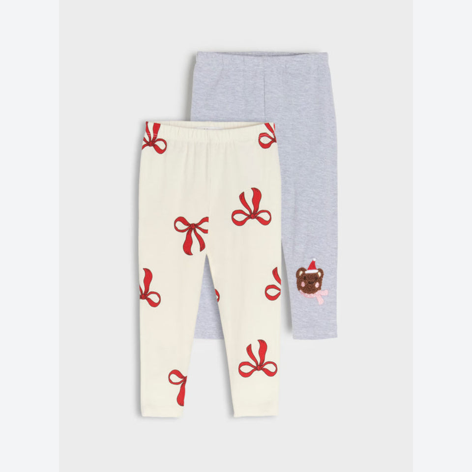 Two pairs of children's leggings laid flat on a light gray background. The front pair is cream with a red bow pattern, and the back pair is light heather gray with a small brown bear graphic.
