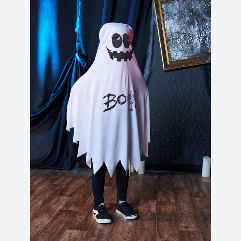 A child wearing a white ghost costume with black jagged hem and a spooky face on the hood, standing on a wooden floor in front of dark draped curtains.
