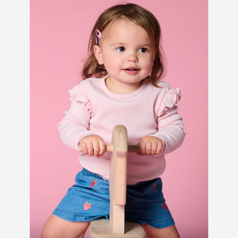 A toddler girl with light brown hair wearing a light pink long-sleeve ruffle-sleeve top and blue denim shorts with pink strawberry details, riding a small wooden toy on a pink background.