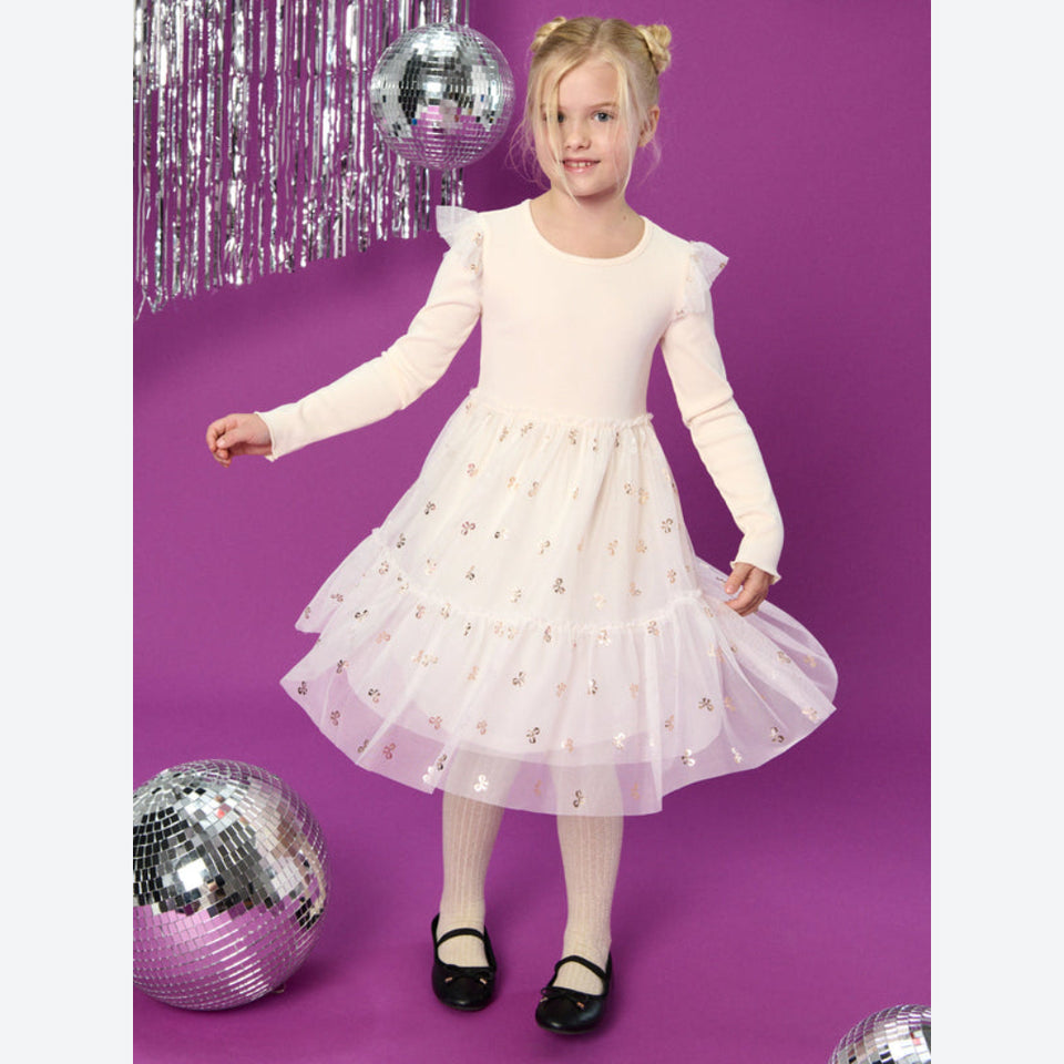 A young girl with blonde hair in a ponytail stands on a purple studio background wearing a long-sleeve cream dress with a sparkly tulle skirt. She is surrounded by silver disco balls and hanging tinsel.