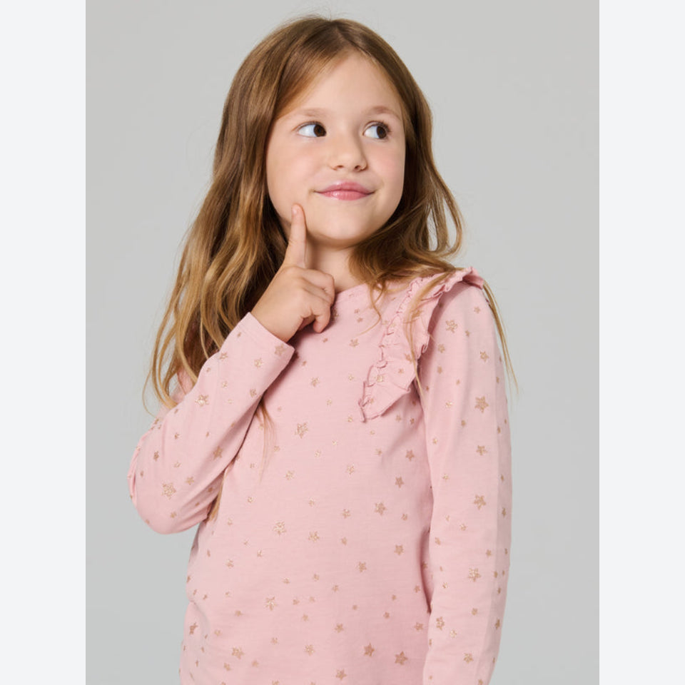 A young girl with long light brown hair wearing a pink long-sleeve top with a gold star pattern, posing against a solid gray background.