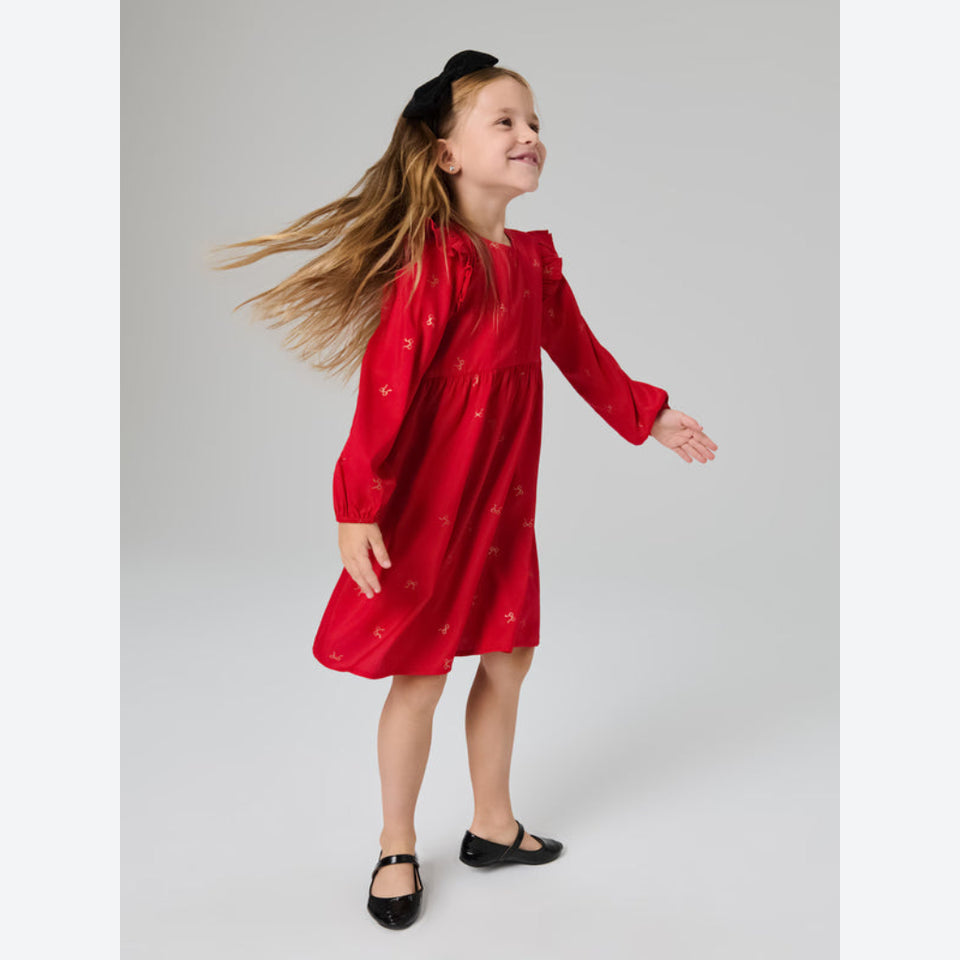 A young girl with long light brown hair wearing a red long-sleeve dress with small gold embroidered motifs, a black velvet bow headband, and black Mary Jane shoes, captured mid-spin against a neutral gray studio background.