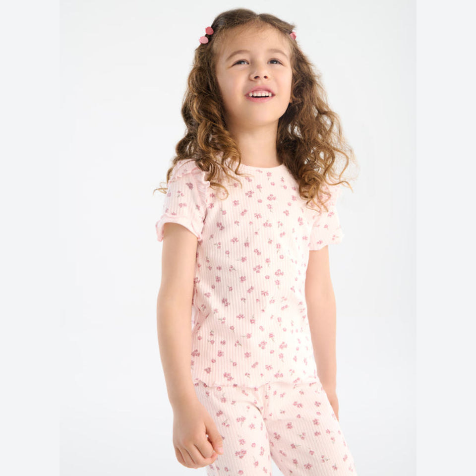 A young girl with curly brown hair wearing a light pink ribbed two-piece pajama set with a small floral print, standing against a plain white background.
